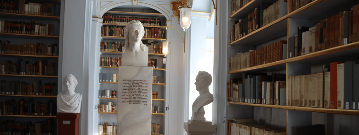 The image shows the ornate Rococo Hall of the historic Duchess Anna Amalia Library in Weimar, Germany, featuring classical busts on pedestals and rows of ancient bookshelves